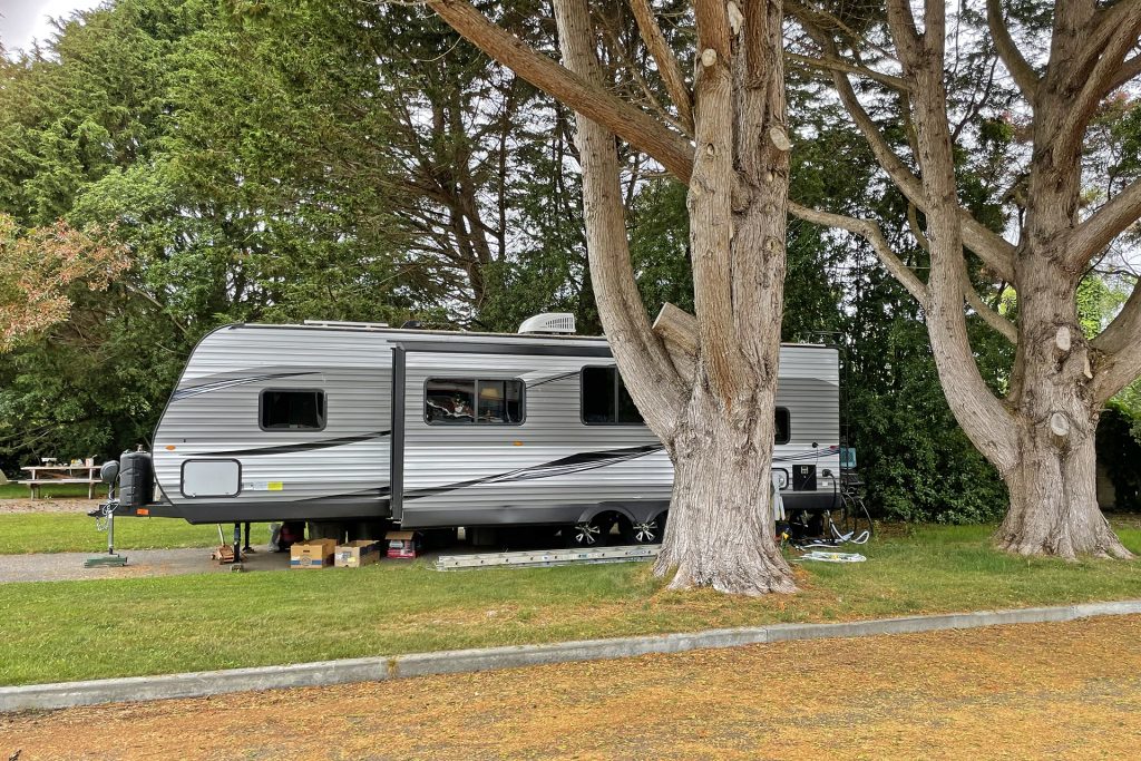 Rv parked underneath several large trees with grassy field